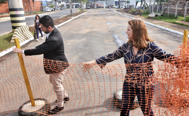 Em uma rua, em pé, o secretário Raizer e a prefeita tiram uma tela laranja que obstrui a passagem de veículos