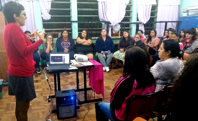 Em uma sala, uma mulher em pé e um grupo de pessoas sentadas a sua frente
