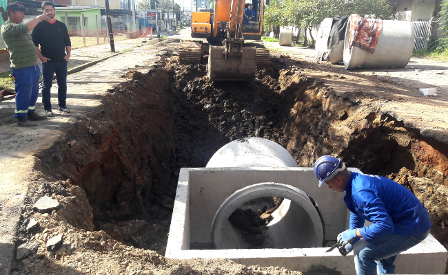 Um grande buraco, com uma caixa de concreto e um cano. Um funcionário está nesta caixa. Ao fundo, cavando um buraco, uma retroescavadeira
