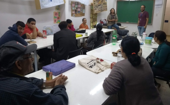 Em uma sala de aula, um homem e uma mulher em pé ao fundo, com alunos sentados no entorno de uma mesa