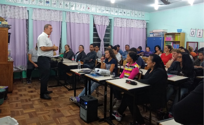 Professor em pé em frente à turma, que está sentada em cadeiras