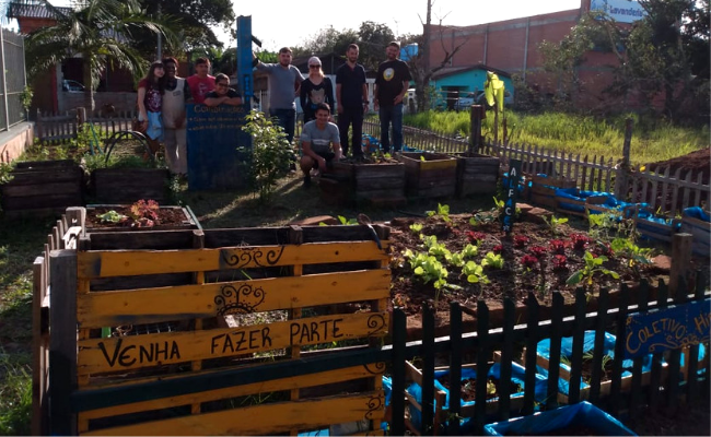 No fundo, um grupo de pessoas lado a lado. À frente, uma horta de plantas e hortas