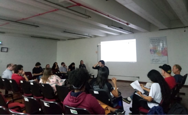 Em uma sala, grupo de pessoas sentadas em cadeiras organizadas no formato de um quadrado