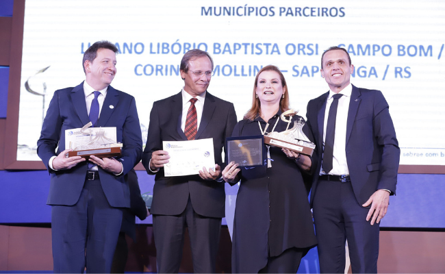 Prefeita em pé em cima de uma palco segurando o troféu. Ao lado, esquerdo dela, dois homens, e a direita um.