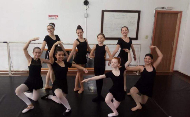 Oito bailarinas vestindo preto em divididas em duas filas de quatro, uma fila em frente a outra, com as bailarinas fazendo "pose"