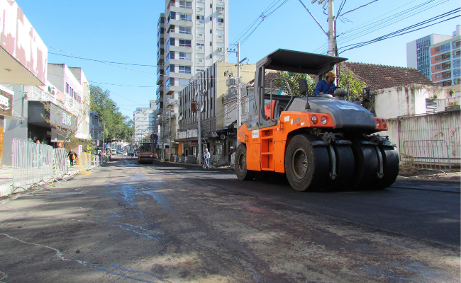 Em uma rua, uma grande máquina com seis pneus, nivelando umaa fina camada de asfalto na via 