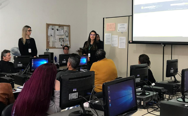 Em uma sala com computadores, duas mulheres em pé ao fundo e alunos sentados nos computadores