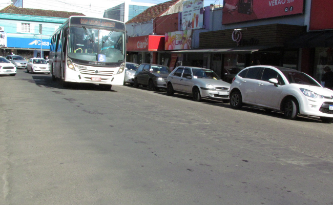 Em uma rua asfaltada e com lojas, um ônibus circula na via