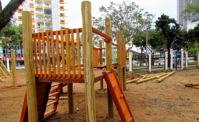 Em uma área com areia no chão, no centro, um brinquedo de madeira no fortmato de uma caixa instalado