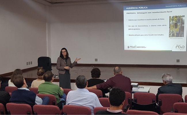 Em um auditório, público sentado e a secretária Roberta à frente em pé