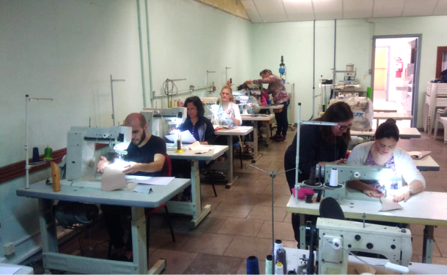 Em uma sala, divididas em duas filas, máquinas de costuras com aprendizes manuseando-as