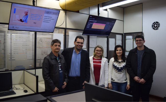 Em uma sala, com dois televisores de fundo, cinco pessoas paradas lado a lado
