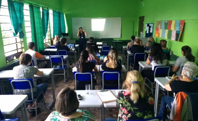 Em uma sala de aula, alunos sentados nas classes e um professor em pé à frente
