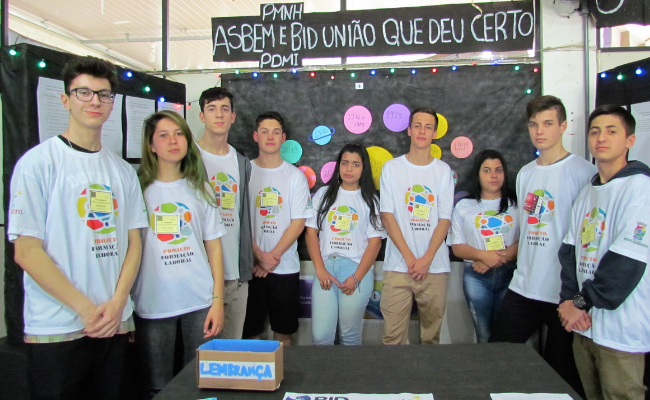 Grupo de alunos em pé vestindo uma camiseta branca dentro do estande na feira