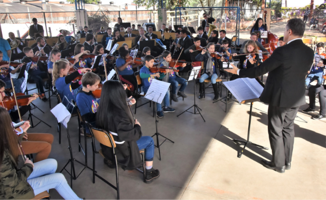 Na quadra da escola, sentados, as orquestras com os instrumentos e o regente à frente