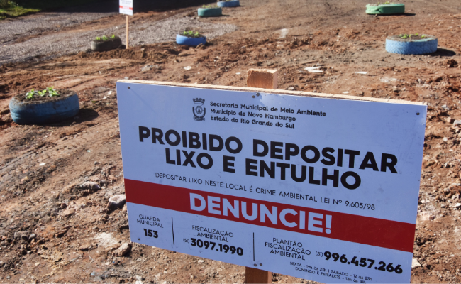 Uma placa branca afixada em um terreno, com pneus com flores plantadas dentro atrás