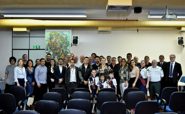 Em um auditório, com as cadeiras à frente, grupo de pessoas lado a lado posam para a foto