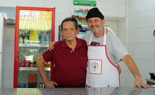 Dois homens abraçados atrás de um balcão dentro de uma banca