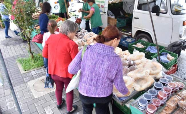 Estande da feira com frutas e verduras expostas e pessoas comprando. Atrás uma kombi