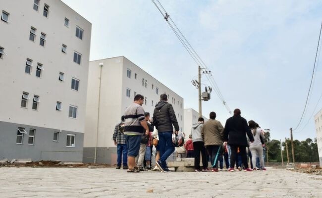 À esquerda, dois grande prédios lado a lado. No centro, grupo de pessoas caminhando de costas