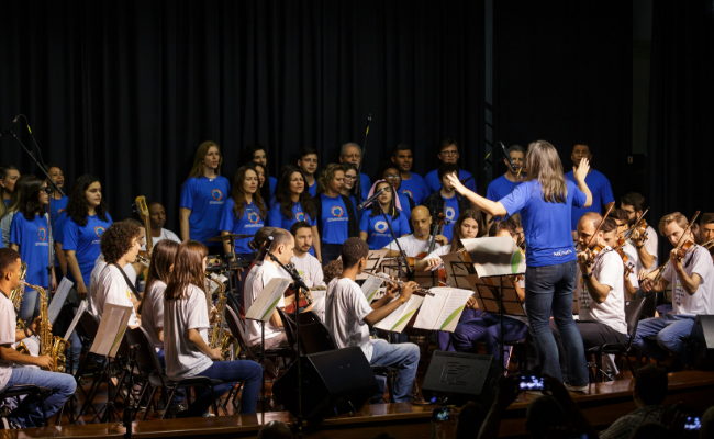 Em um palco, alunos com os instrumentos musicais e o coral se apresentam