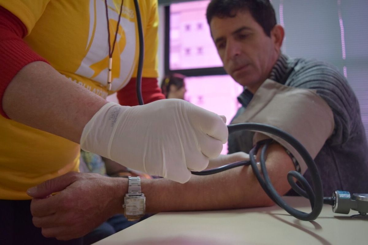Um menino sentado e um enfermeira em pé. Ela, com um aparelho no braço dele, mede a sua pressão arterial