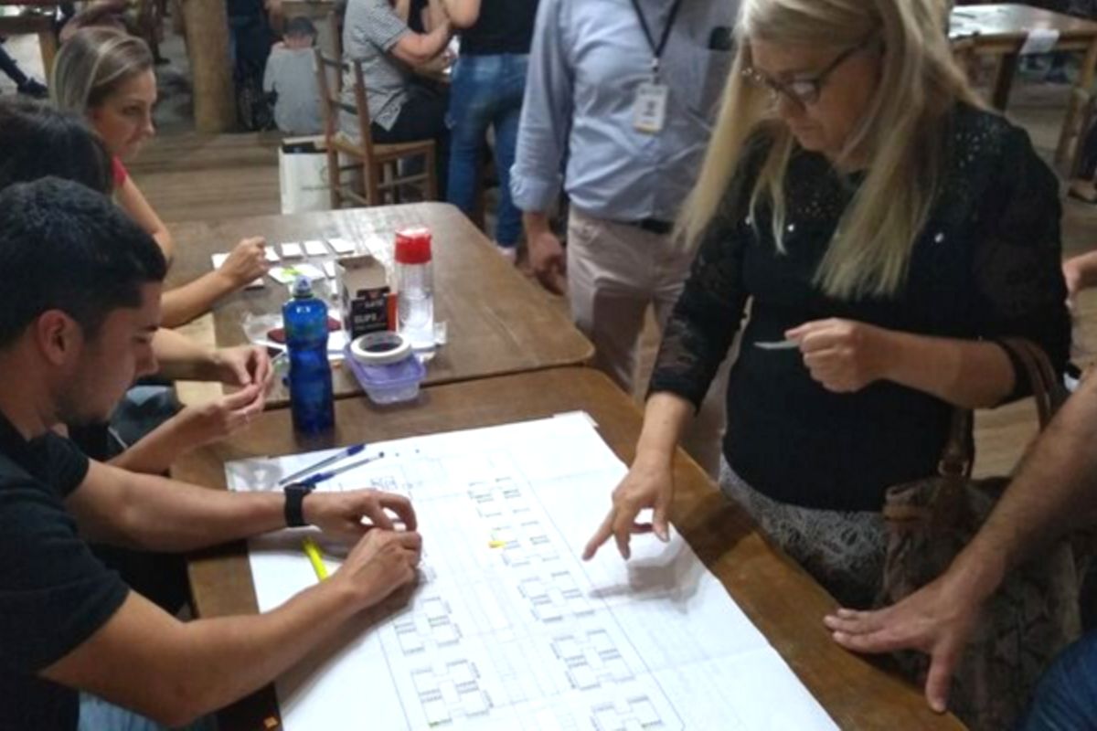 Em uma mesa, à esquerda, duas pessoas sentadas e, do outro lado, uma mulher em pé apontando para uma folha de papel