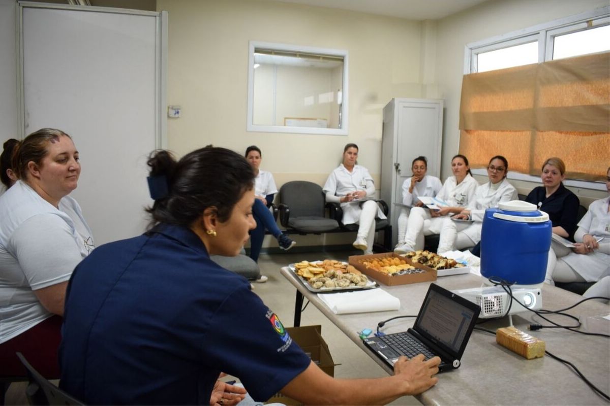 Em uma sala, mulheres sentadas vestindo um jaleco branco e uma, em um computador, de azul