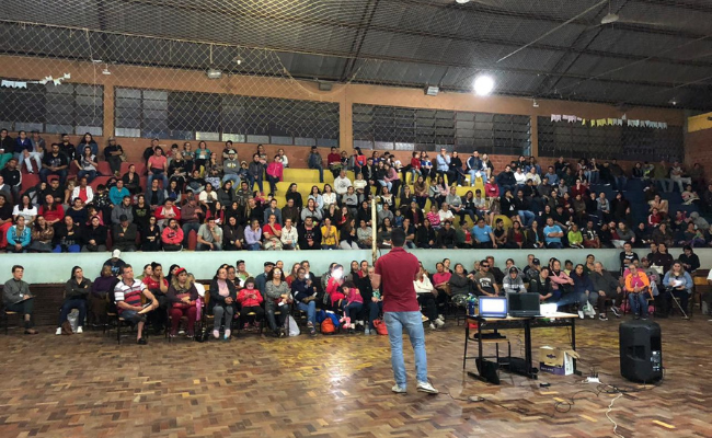 Ao fundo, uma arquibancada com pessoas sentadas. A sua frente, um homem em pé ao microfone