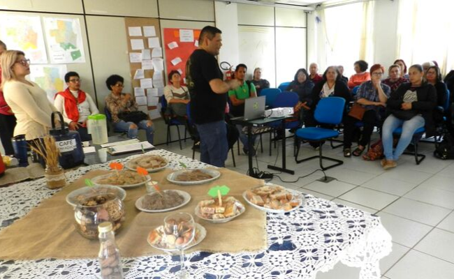Em uma sala, à esquerda uma mesa cheia de comida. No centro, um homem em pé falando para uma platéia sentada à direita