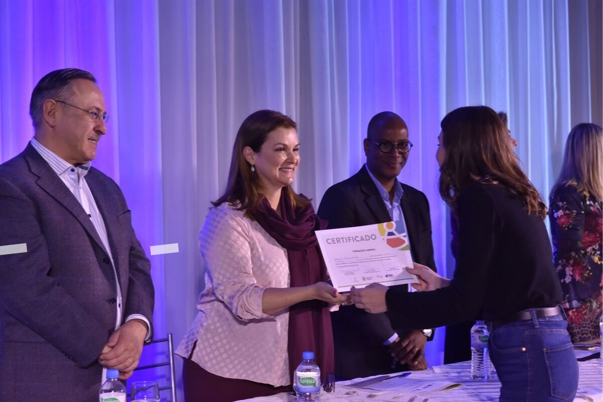 Prefeita Fátima, atrás de uma mesa, entregando um diploma a uma aluno, que está no outro lado da mesa