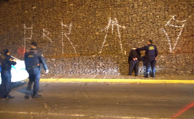 Ao lado de um grande muro de pedras, guarda municipal segura homem que estava pichando