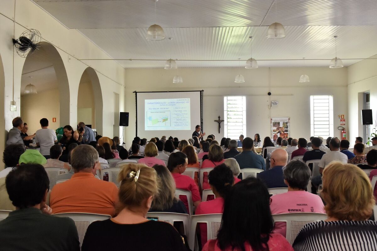 Homem ao microfone ao fundo e pessoas sentadas em cadeiras