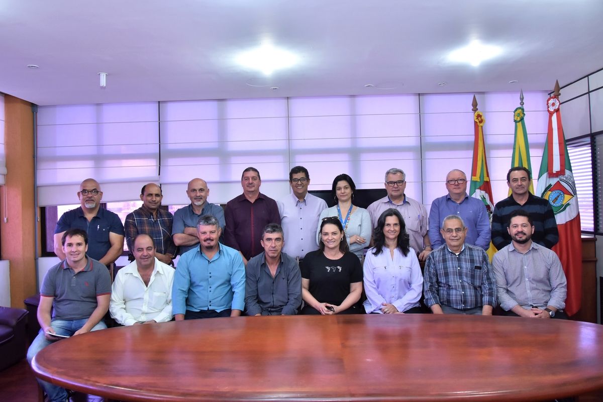 Grupo de pessoas sentadas lado a lado e outro grupo de pessoas em pé atrás desta pessoas. Os dois grupos estão atrás de uma mesa
