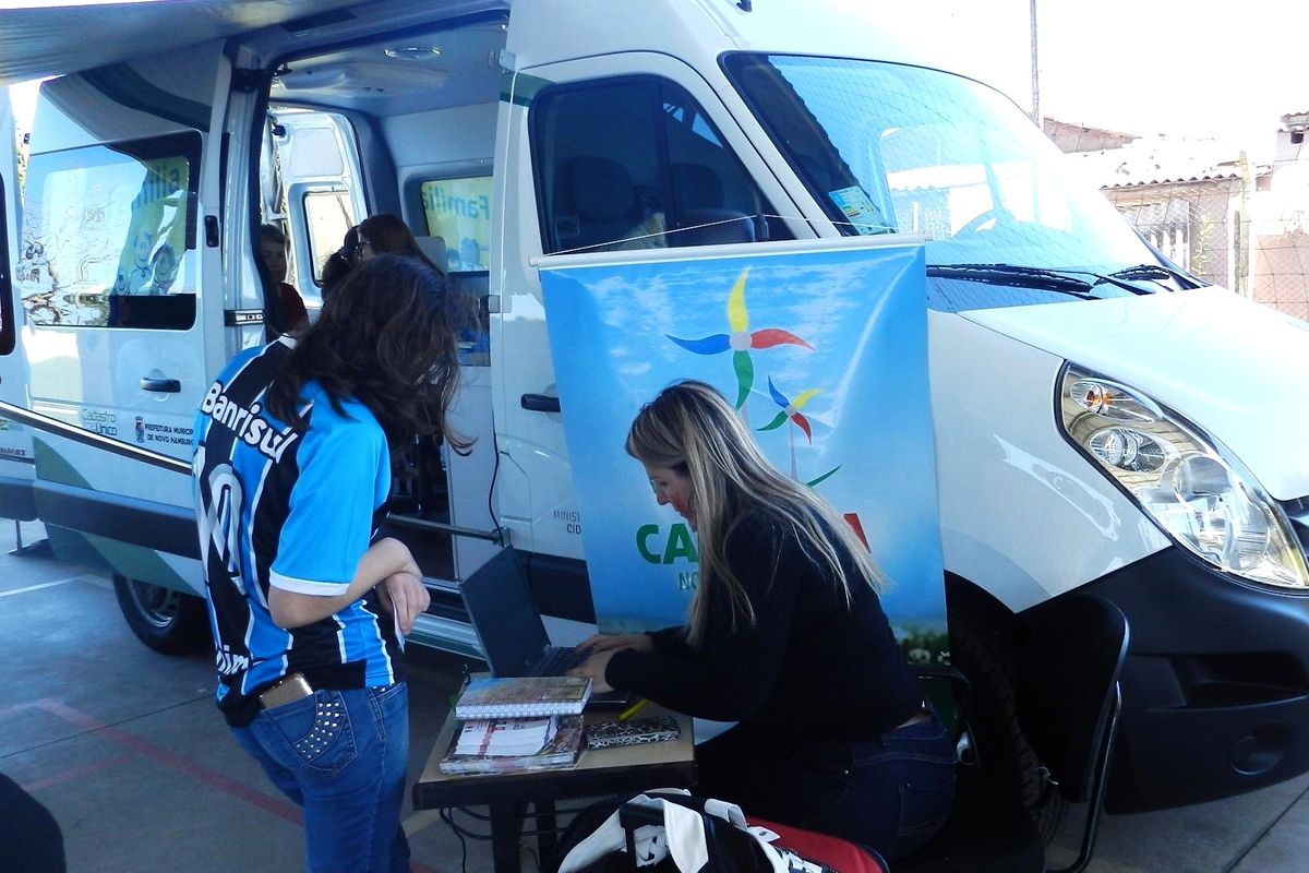 Uma van branca estacionada com uma mulher sentada ao lado em frente a uma mesa. Uma mulher em pé é atendida pela mulher sentada