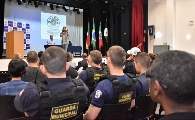 Em um auditório, ao fundo, prefeita em pé ao microfone em um palco, e guardas municipais sentados