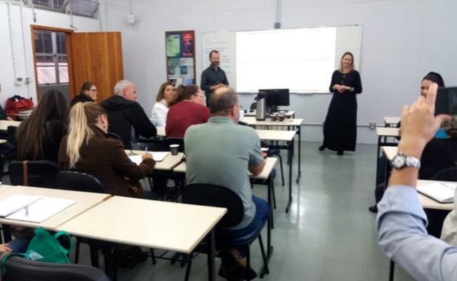 Em uma sala, pessoas sentadas em classes escolares e o diretor Deivid em pé ao fundo