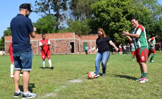 Em um campo de futebol, prefeita chutando uma bola com jogadores no seu entorno