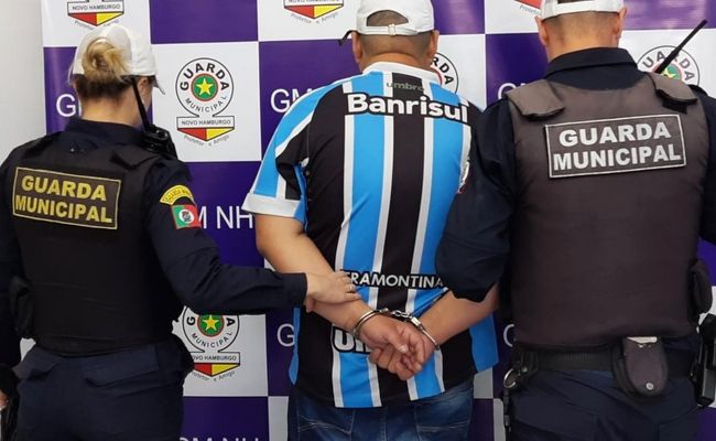 Três pessoas de costas, sendo um homem algemado com a camisa do time do Grêmio no meio, e dois guarda municipais fardados nos lados, sendo uma mulher à esquerda e a direita um homem