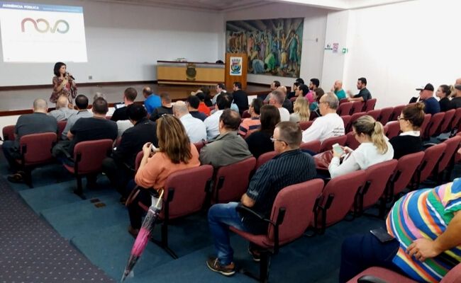 Secretária Roberta ao fundo em pé ao microfone e público sentado em cadeiras