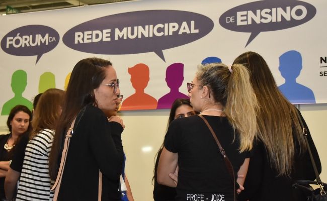Mulheres em pé e um mural ao fundo escrito Fórum da Rede Municipal de Ensino