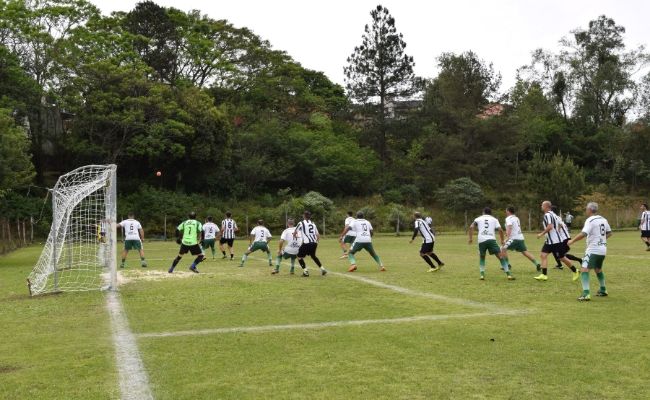 Em um campo de futebol, um time de branco contra um time de verde durante a cobrança de um escanteio. A grande área está cheia de jogadores e a bola no ar vindo em direção à áerea