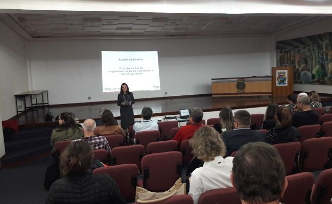 Em um auditório, público sentado e a secretária Roberta à frente em pé