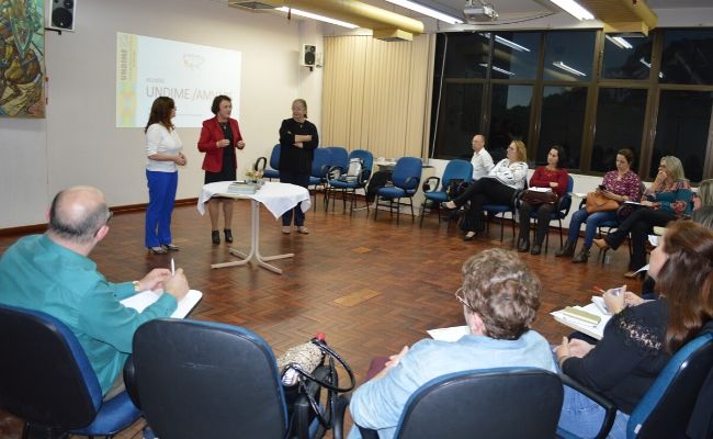 Em formato de "U", pessoas sentadas em cadeiras e três mulheres em pé no centro