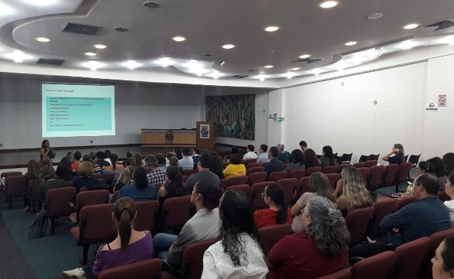 Em um auditório, público sentado e mulher ao microfone ao fundo em pé
