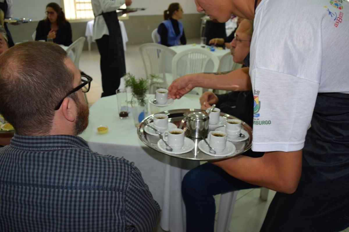 Jovem em pé ao lado de uma mesa com uma bandeja na mão. Dois homens sentados em cadeiras junto à mesa