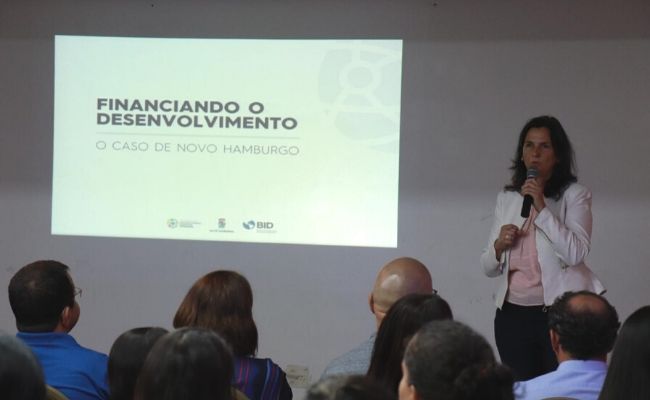 Secretária Paraskevi em pé ao microfone. Nas suas costas é projetada uma tela na parede. A sua frente, pessoas sentadas em cadeiras