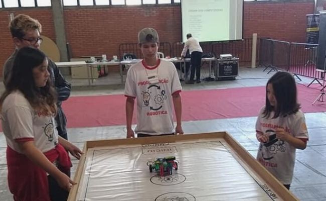 Crianças no entorno de uma mesa, com um robô em formato de carro sendo movimentado em cima desta mesa