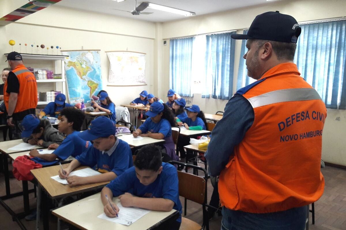 Em uma sala de aula, crianças sentadas nas classes e homem em pé de colete laranja escrito defesa civil