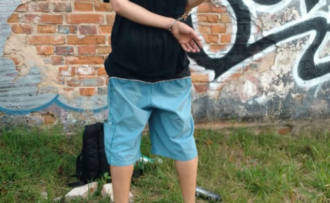 Homem de costas com as mãos para atrás algemada e um mochila e latas de tinta no chão